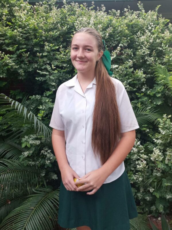 A young woman with long hair stands smiling in front of lush greenery. She wears a white blouse and green skirt, exuding a cheerful, relaxed demeanor.
