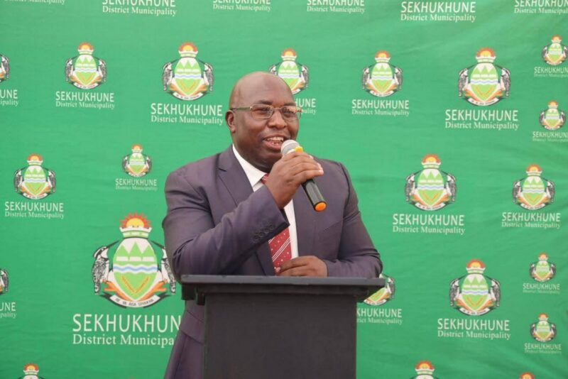 A man in a suit speaks into a microphone at a podium. The backdrop displays the Sekhukhune District Municipality logo repeatedly on a green background.
