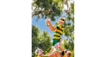 Rugby player in green and yellow is lifted to catch the ball during a game. Teammates support him with focus and teamwork under clear skies.