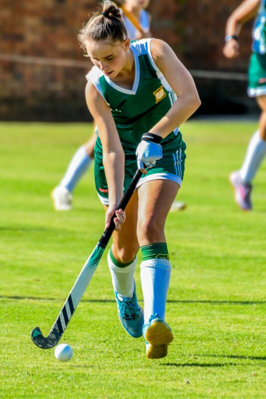A field hockey player in a green and white uniform focuses intensely as she dribbles the ball with her hockey stick on a grassy field.