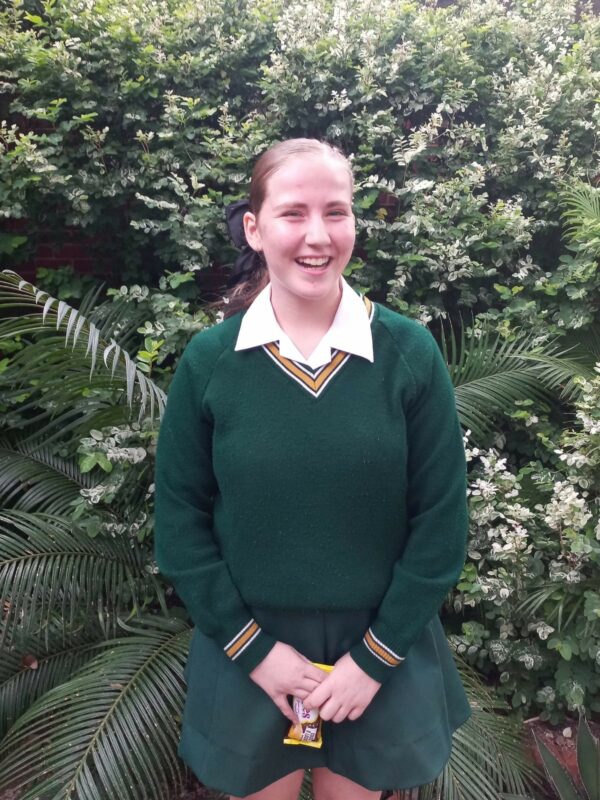 A person with a joyful expression stands in front of lush green foliage, wearing a green sweater and skirt. They hold a small yellow object.