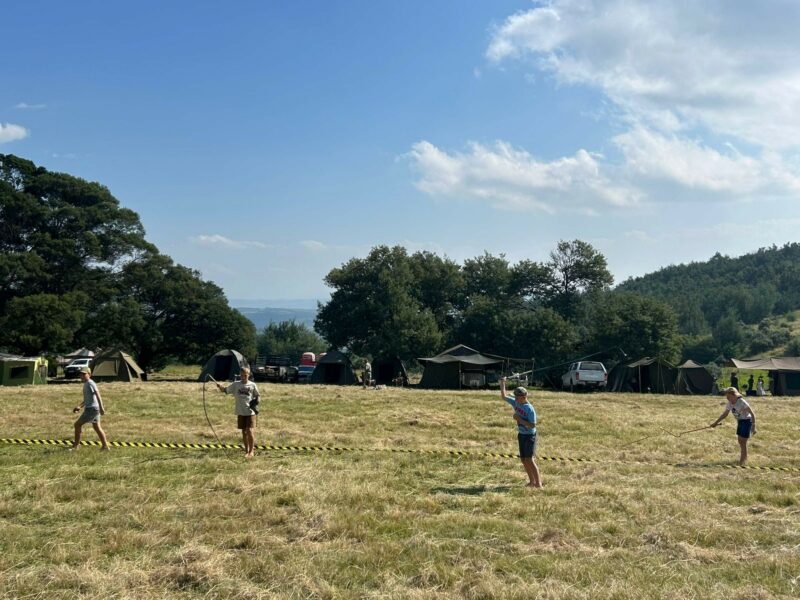 Four people participate in an outdoor activity on a sunny field near tents and trees. The scene exudes a lively, adventurous camping vibe.