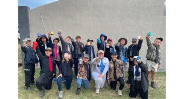 A group of children stands cheerfully in front of a plain wall, holding colorful mugs aloft. They are dressed in casual outdoor clothing, conveying excitement.