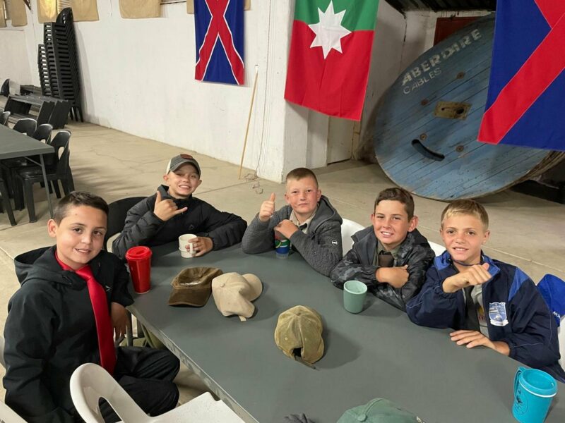 Five young boys sit at a table, smiling and giving thumbs up. They are dressed casually, surrounded by colorful flags in a bright, spacious room.