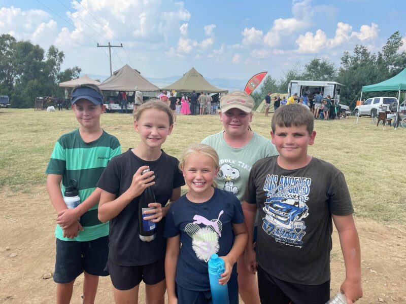 Five smiling children stand outdoors on a sunny day, holding water bottles. Tents and people are visible in the background, creating a lively atmosphere.