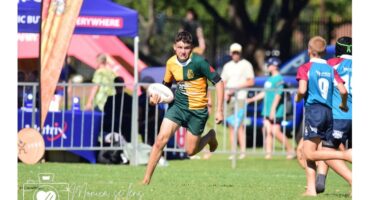 A young rugby player in green and yellow runs with the ball on a grassy field, focused on the game. Teammates and spectators are visible in the background.