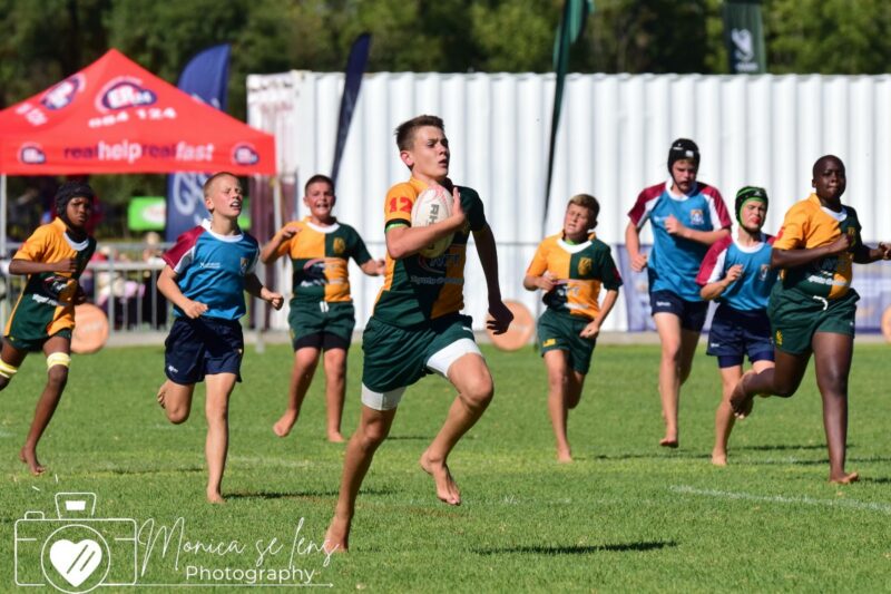 A young rugby player in a green and orange uniform sprints across a grass field, clutching the ball. Teammates and opponents chase him under a bright sky.