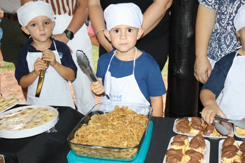 Two children wearing chef hats and aprons stand at a food stall. They hold tongs near trays of fried noodles, pastries, and covered desserts. The scene is lively and engaging.