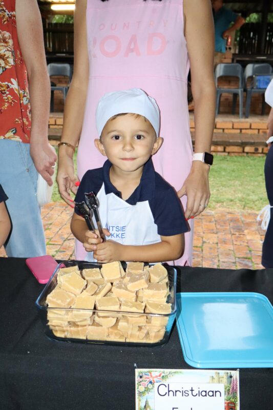 A young child wearing a chef hat and apron holds tongs next to a tray of fudge at a table, smiling, with adults standing nearby.