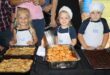 Three children wearing aprons and chef hats stand behind trays of food, including stuffed pasta and desserts, smiling proudly at a cooking event.