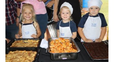 Three children wearing aprons and chef hats stand behind trays of food, including stuffed pasta and desserts, smiling proudly at a cooking event.