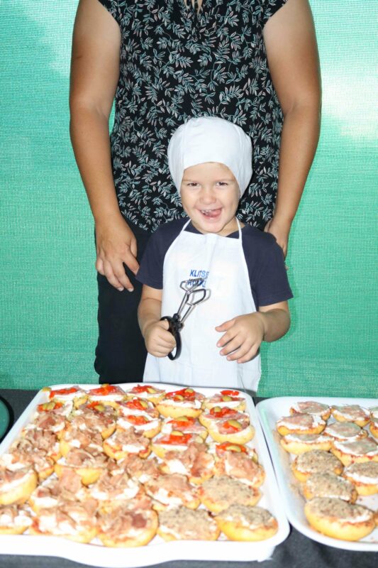 A smiling child in a chef's hat and apron holds a whisk, standing in front of trays of mini pizzas. An adult stands behind, hands gently on the child's shoulders.