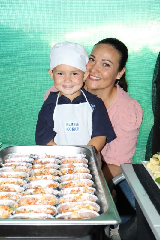 A smiling child in a chef uniform stands beside a tray of baked goods in foil cups; a woman crouches beside them, smiling warmly. Bright, joyful atmosphere.
