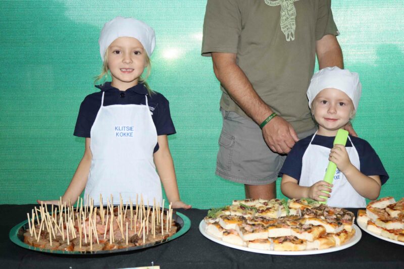 Two young children in white chef hats and aprons stand smiling behind a table with skewered snacks and pizzas. An adult stands next to them, creating a cheerful atmosphere.