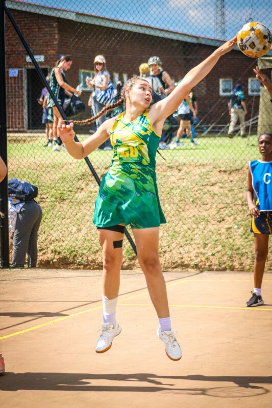 A female netball player in a green and yellow uniform leaps to intercept the ball. Background shows a blurred fence and spectators, conveying action and focus.