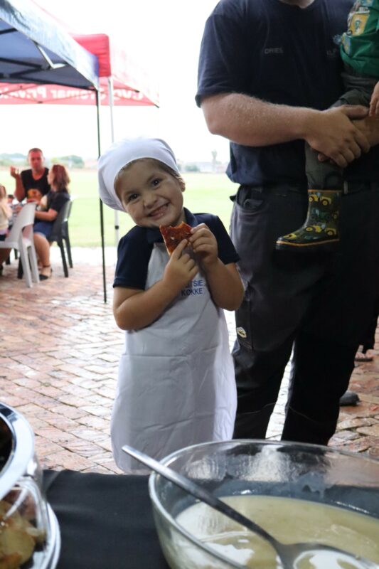 A young child in a chef's hat and apron smiles while holding a slice of pizza. The atmosphere is cheerful under an outdoor canopy.