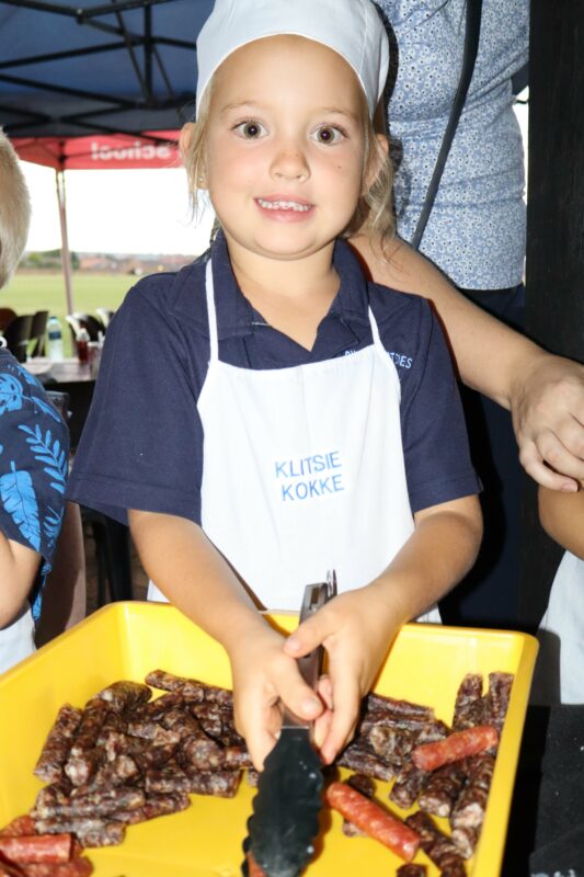 A young child in a white chef's hat and apron holds tongs over a yellow tray filled with sausages. The atmosphere is lively and cheerful.
