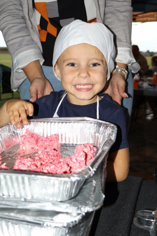Young child in a chef hat and apron smiles while handling pink dough in aluminum trays, conveying joy and creativity in a cooking activity.