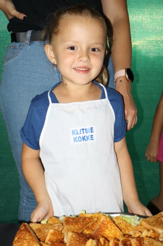 Young girl in a white apron with "KLITSIE KOKKE" text, smiling while standing in front of a table filled with toasted sandwiches. Bright, cheerful atmosphere.