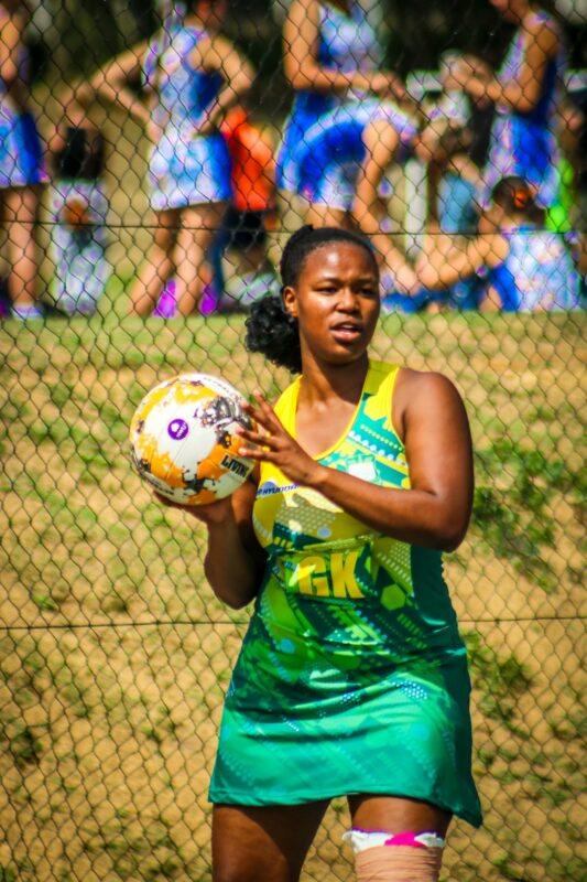 A netball player in a green and yellow uniform holds a ball, focusing intensely. Teammates and spectators are blurred in the sunny background.