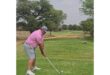 A golfer in a pink shirt and gray shorts prepares to swing on a lush, green fairway. Trees line the course under a slightly overcast sky.