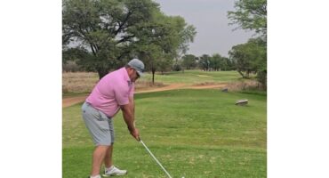 A golfer in a pink shirt and gray shorts prepares to swing on a lush, green fairway. Trees line the course under a slightly overcast sky.