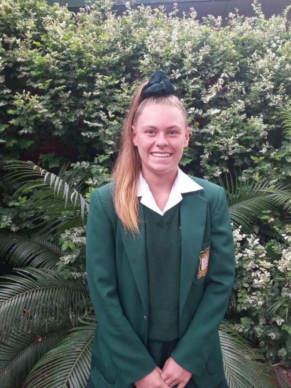 A smiling person in a green school uniform stands in front of lush greenery. The tone is cheerful and the setting appears to be a garden.