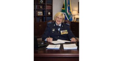 An official in a navy blue uniform with medals sits smiling at a wooden desk, pen in hand, amidst paperwork. Flags and a bookshelf are in the background.