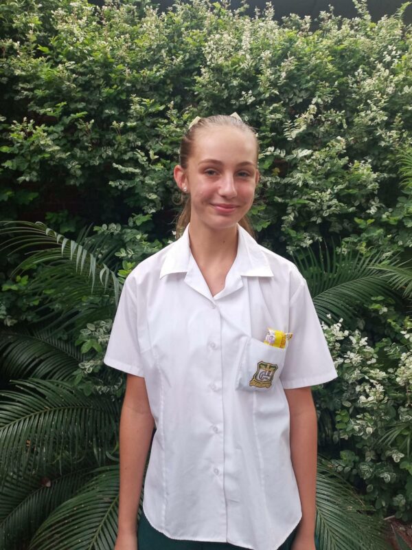 A young girl in a white school uniform with a badge smiles softly, standing in front of lush green bushes. The scene is bright and serene.