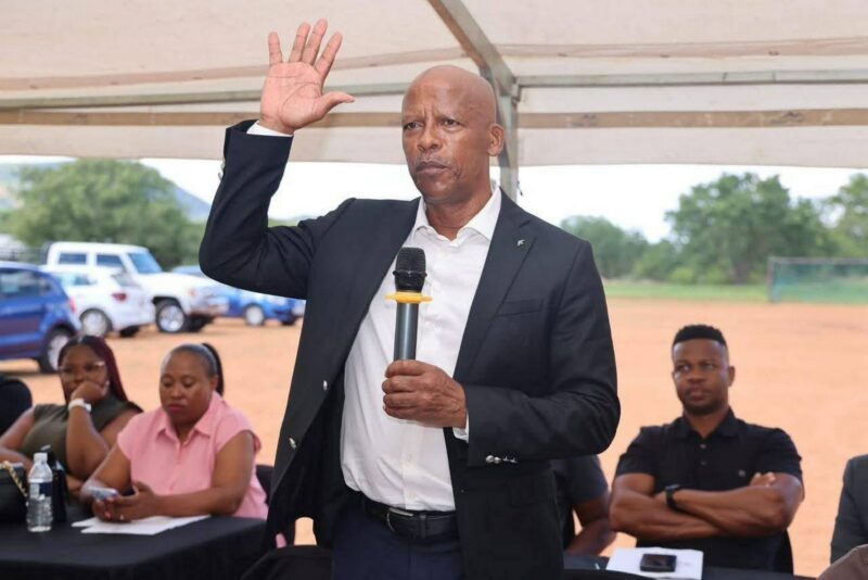 A man in a suit holds a microphone and raises his hand, speaking under a tent. Seated people listen attentively. The setting is outdoors with parked cars.