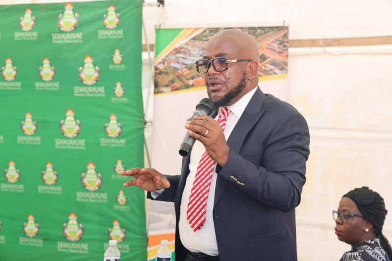Man in a suit and striped tie speaks into a microphone at an event. A green banner with logos is in the background. The setting is professional.