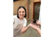 A smiling woman in a white shirt holds a large tarantula with another person. The background is a brick-patterned wall, creating a relaxed, adventurous mood.