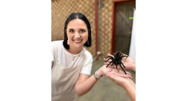A smiling woman in a white shirt holds a large tarantula with another person. The background is a brick-patterned wall, creating a relaxed, adventurous mood.