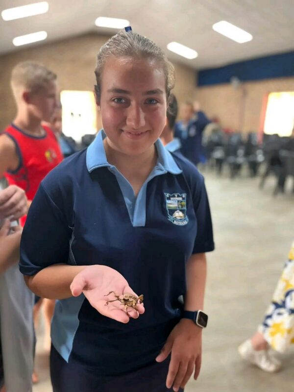 A person in a navy blue school uniform holds a scorpion in their palm, smiling confidently. The background shows a spacious room with other people.