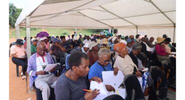 A group of people sit attentively under a large white tent, engaged in a document review. The scene conveys concentration and participation.