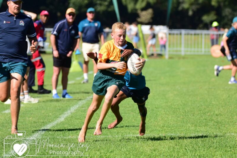 Young rugby players on a field, one in a yellow jersey clutching a ball as another tackles him. Coaches and spectators are visible in the background.