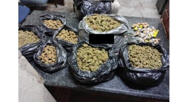 Numerous black bags filled with marijuana buds and smaller packages of various items are spread across a dark, textured table indoors.