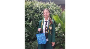 A young person in a green school uniform with badges stands smiling, holding a blue gift bag. They're in front of lush green bushes, conveying a sense of achievement and celebration.