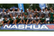 A rugby team poses together, wearing gray and green uniforms, behind a large blue "Nashua" banner. The setting is outdoors with trees behind. The mood is cheerful.