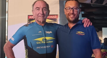 Two men stand smiling at a cafe entrance. One wears a blue cycling kit, the other a blue polo. The atmosphere is warm and friendly, with people and bicycles in the background.