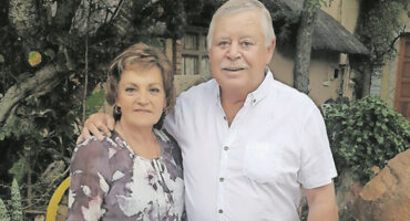Elderly couple smiling warmly, standing outdoors. The man in a white shirt, arm around the woman in a purple floral blouse. Lush greenery surrounds them.