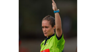 A referee in a bright green shirt raises one arm, possibly signaling in a sports event. The expression is focused, suggesting concentration.