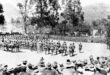 A historical black-and-white photo shows a large assembly of soldiers in uniform gathered under tall trees. A crowd watches in the foreground. War atmosphere.