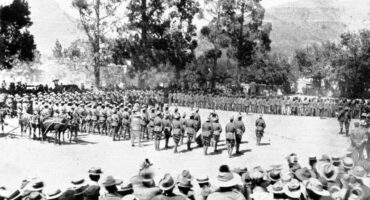 A historical black-and-white photo shows a large assembly of soldiers in uniform gathered under tall trees. A crowd watches in the foreground. War atmosphere.