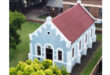 Aerial view of a small blue church with white trim and a red roof, surrounded by green lawns and trees, conveying a serene and historic ambiance.