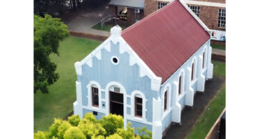 Aerial view of a small blue church with white trim and a red roof, surrounded by green lawns and trees, conveying a serene and historic ambiance.