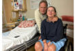 A man and woman pose in a hospital room. The woman is in a wheelchair, wearing a navy top and blue shorts. The man stands beside her, embracing her shoulder. They both smile softly. A hospital bed with a patterned pillow and some personal items are in the background, creating a warm and supportive atmosphere.