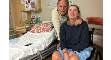 A man and woman pose in a hospital room. The woman is in a wheelchair, wearing a navy top and blue shorts. The man stands beside her, embracing her shoulder. They both smile softly. A hospital bed with a patterned pillow and some personal items are in the background, creating a warm and supportive atmosphere.