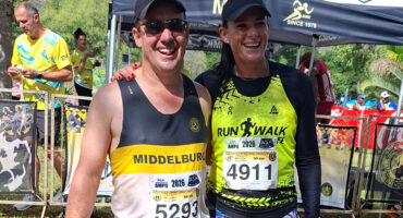 Two smiling runners pose for a photo wearing race bibs, surrounded by other participants and a race tent. The mood is celebratory and energetic.