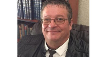 A smiling person wearing glasses and a leather jacket sits on a chair. Books are visible on a bookshelf in the background, creating a cozy setting.